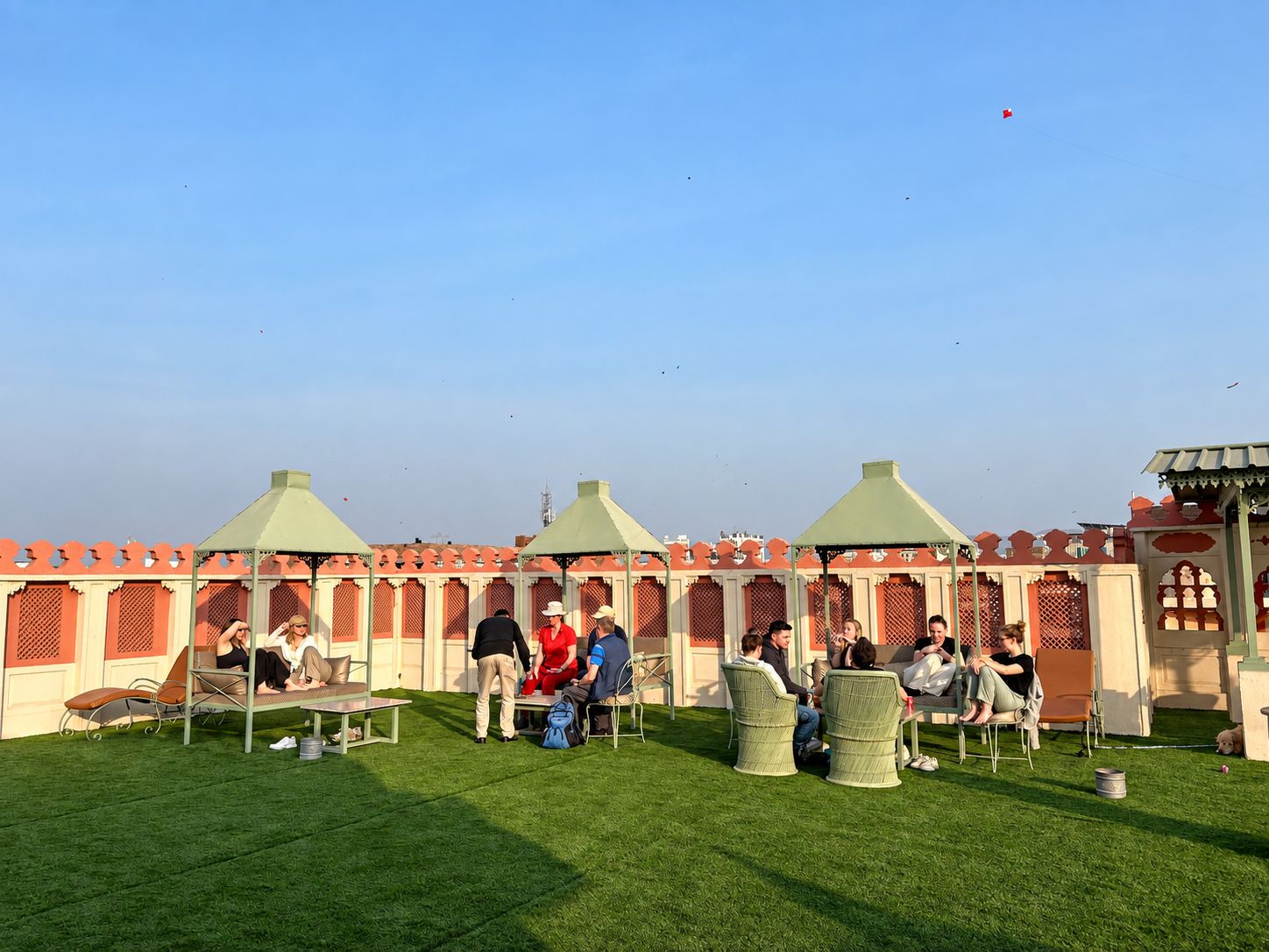 Un espace extérieur aménagé avec des chaises et des tables vertes, entouré d’un bâtiment aux murs rouges et blancs et aux toits verts.