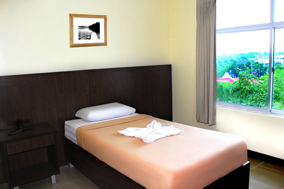 Ein ordentlich bezogenes Bett mit einem weißen Kopfkissen und einem gefalteten Handtuch, ein Nachttisch mit einem Telefon und ein gerahmtes Bild an der Wand.