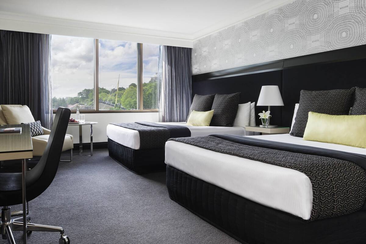 A hotel room with two beds, a desk, a chair, and large windows offering a city view.