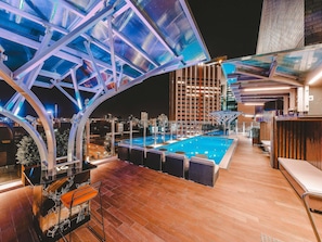 Piscina no terraço com teto de vidro, piso de madeira e área de bar.