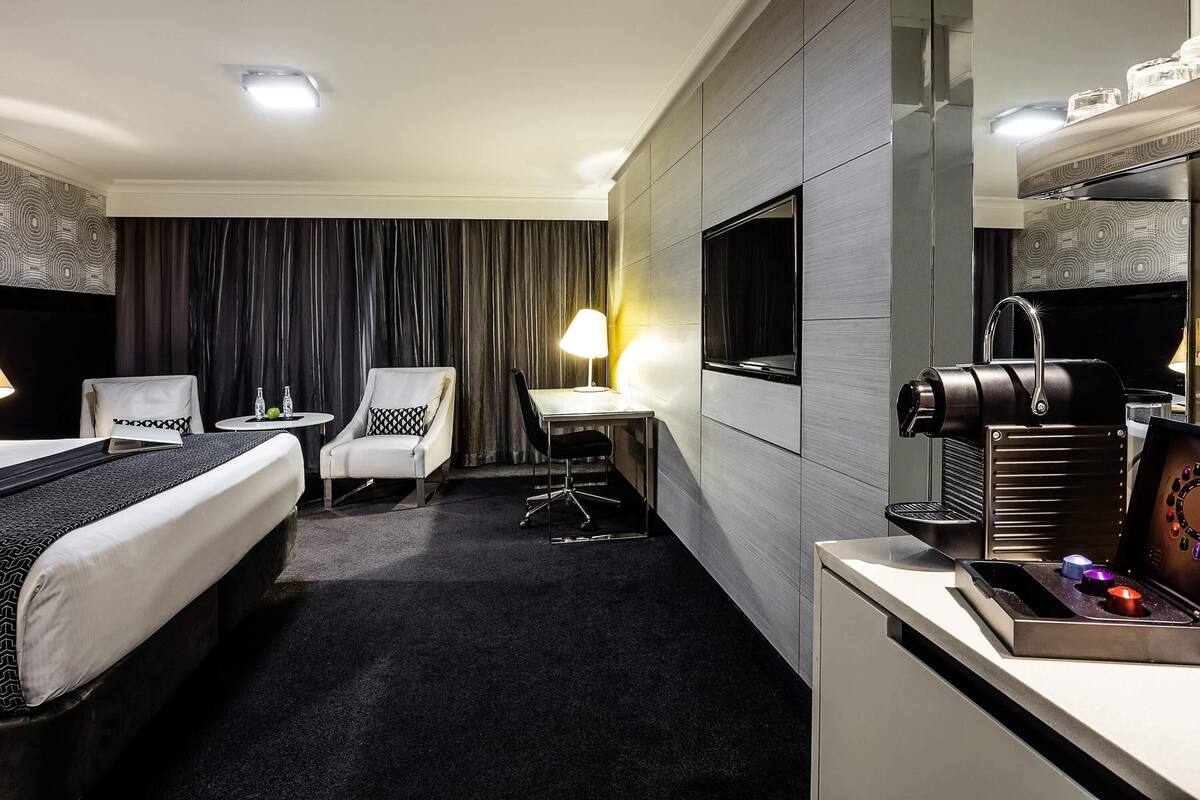 A hotel room with a bed, two chairs, a desk, a TV, and a minibar.