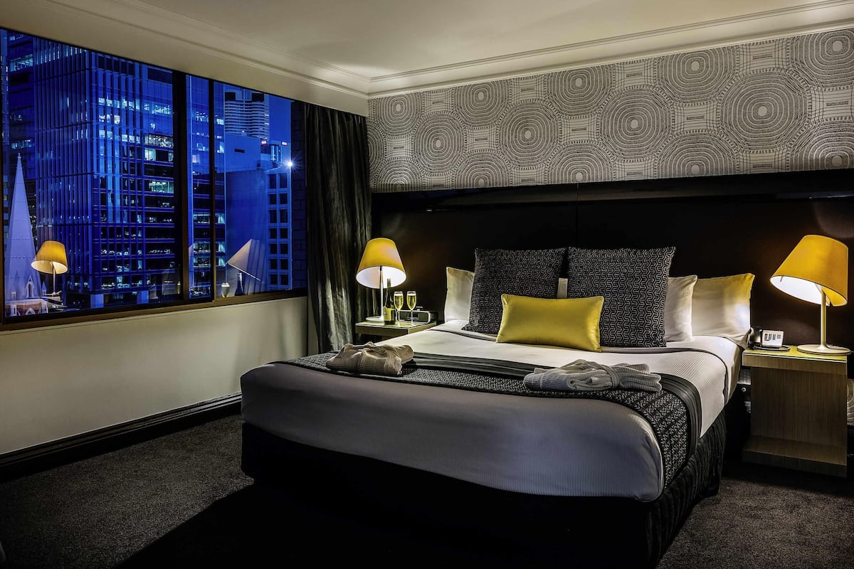 A hotel room with a large bed, two bedside tables, a window with a city view, and patterned wallpaper.