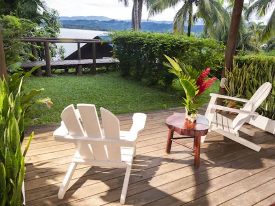 Een terras met twee witte stoelen en een kleine tafel, omringd door weelderige begroeiing en uitzicht op een waterpartij.