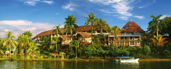 A tropical resort with thatched-roof buildings, palm trees, and a boat on the water.