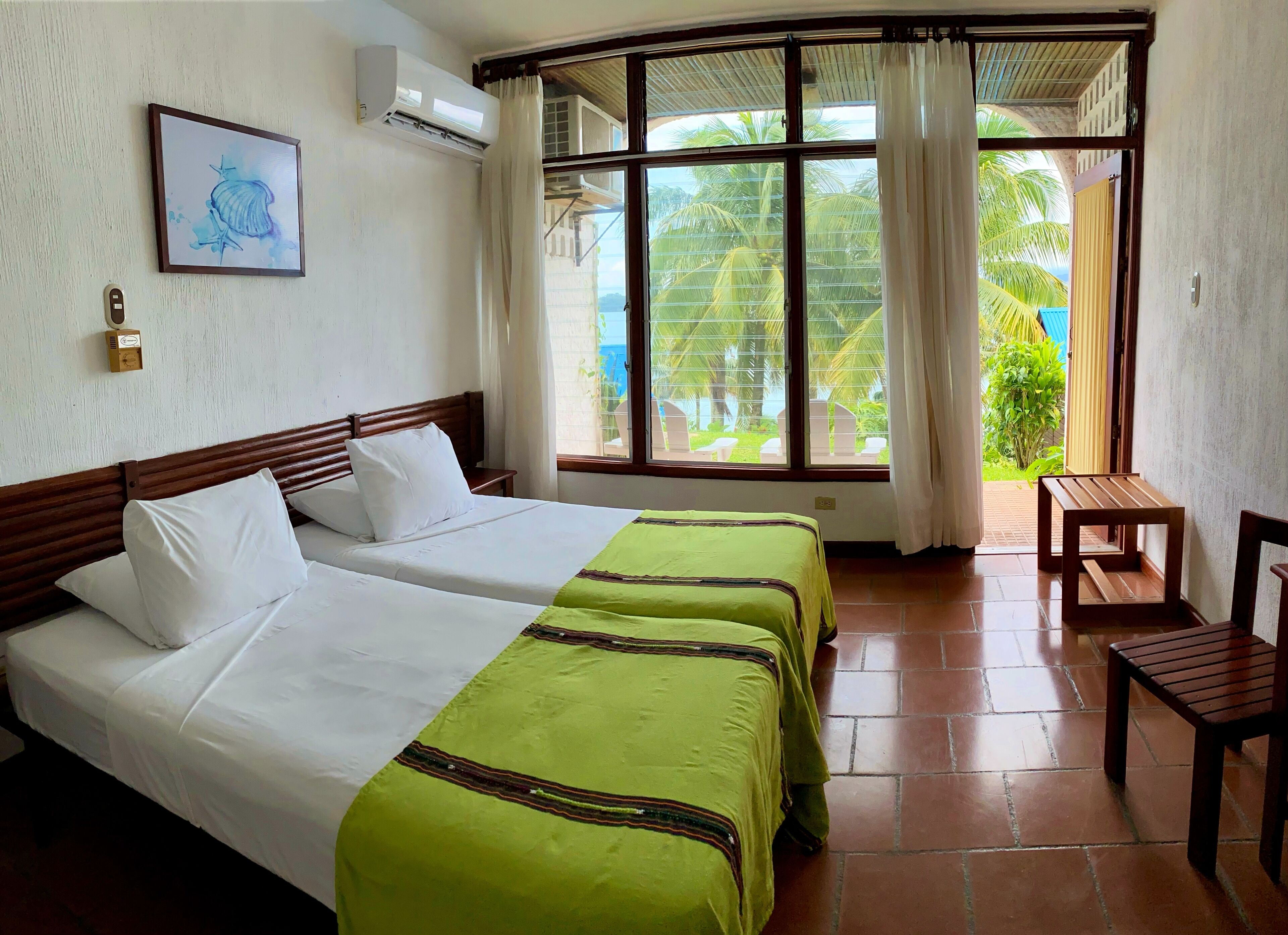 A hotel room with two beds, a wooden headboard, a green bedspread, a small wooden table, a chair, a window with curtains, an air conditioning unit, and a framed picture on the wall.