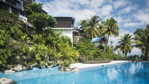 Piscina de resort con agua cristalina azul, rodeada de exuberante follaje y palmeras, con un edificio al fondo.