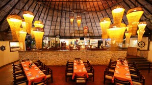 Un restaurante con techo de paja, varias mesas dispuestas con manteles naranjas y lámparas de mimbre colgantes.