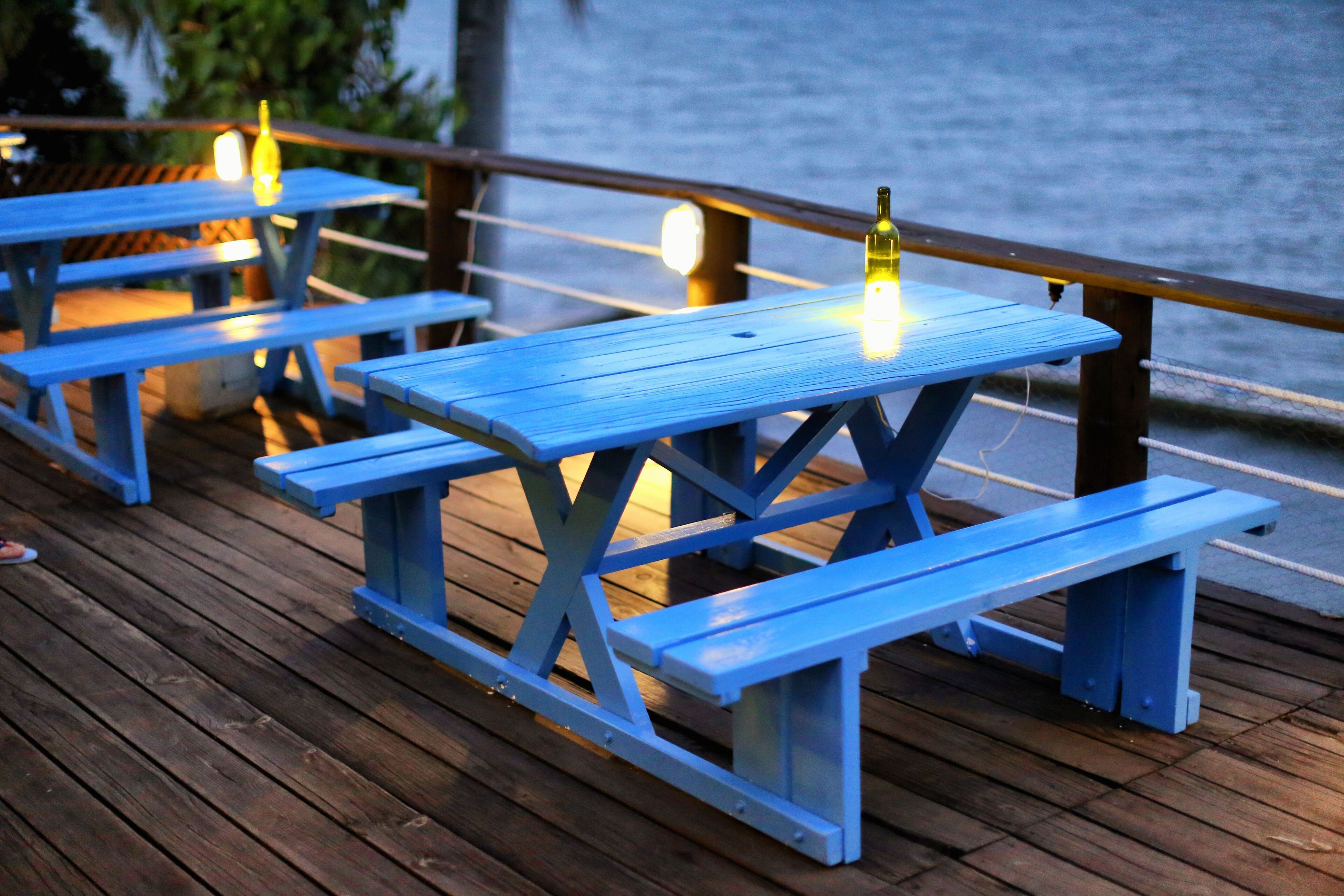Outdoor wooden picnic tables with benches, a bottle of wine, and string lights on a wooden deck by a body of water.