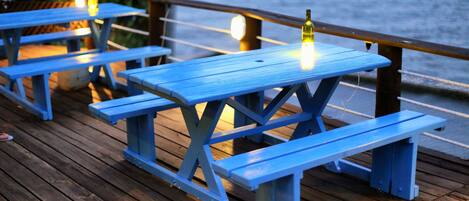 Outdoor wooden picnic tables with benches, a bottle of wine, and string lights on a wooden deck by a body of water.