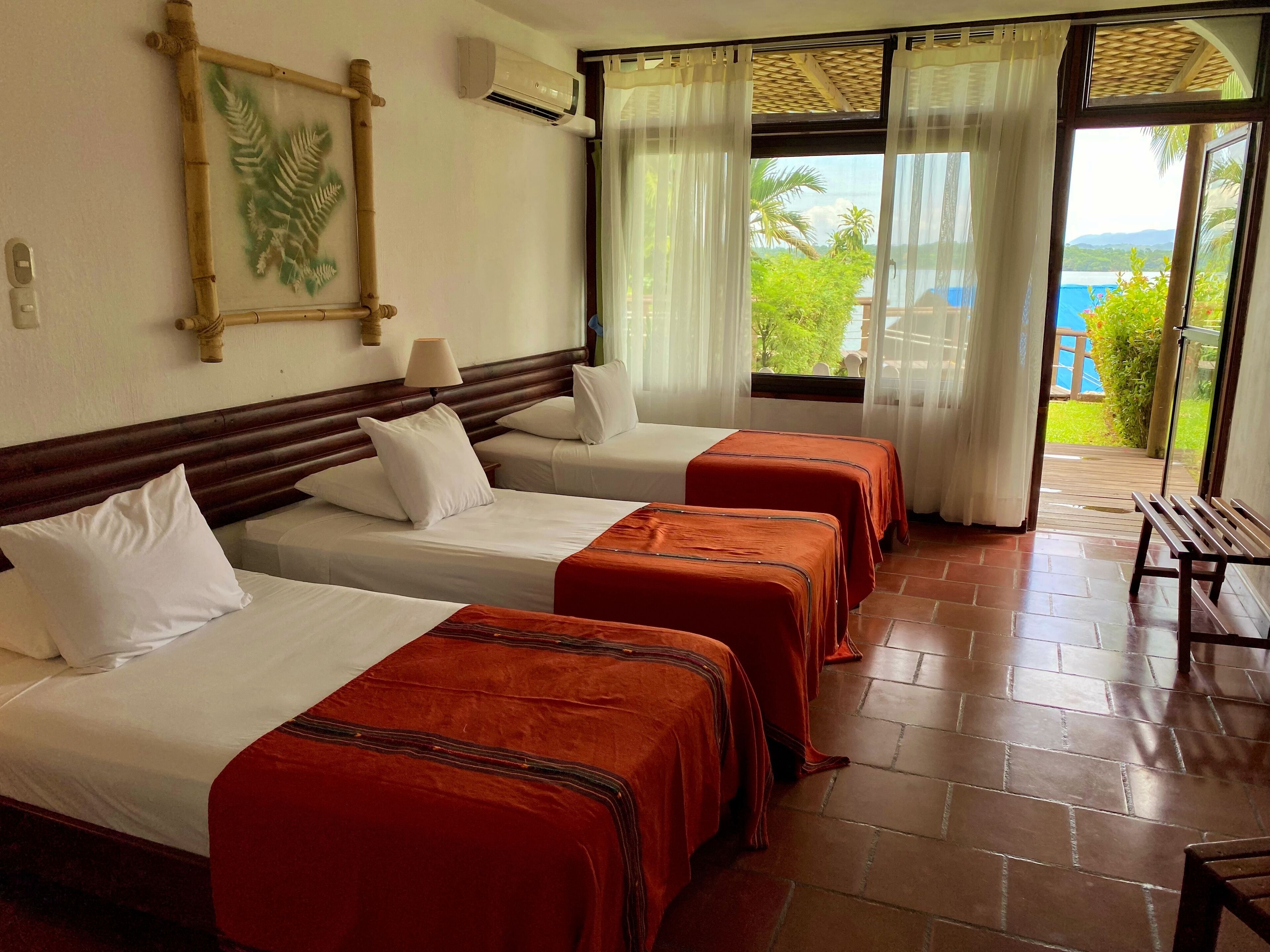 A hotel room with three beds, a wooden headboard, a large window with curtains, and a view of the outdoors.
