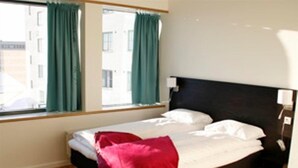 A single bed with a black headboard, white bedding, and a red throw blanket. There are two windows with green curtains, and the room has wooden flooring. - Hotell Montebello (Oslo)