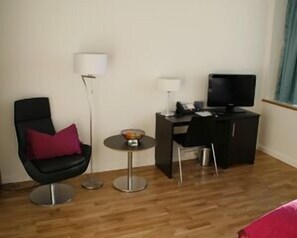 A hotel room with a black chair, a small round table with a bowl, a desk with a computer and a TV, and a lamp. - Hotell Montebello (Oslo)