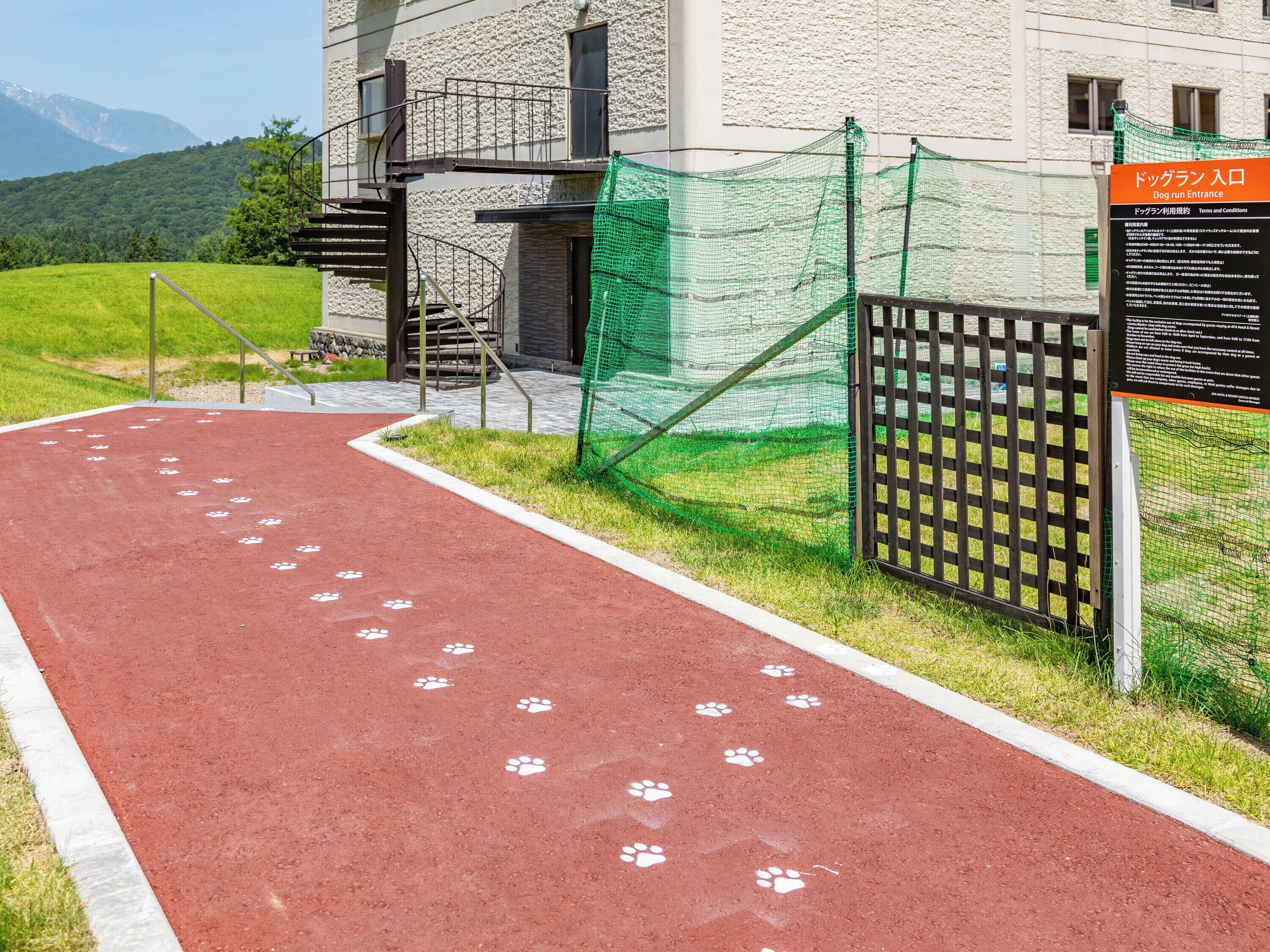 Una pista di atletica rossa con impronte bianche a zampa, un'area recintata verde per cani e un edificio con facciata in pietra.