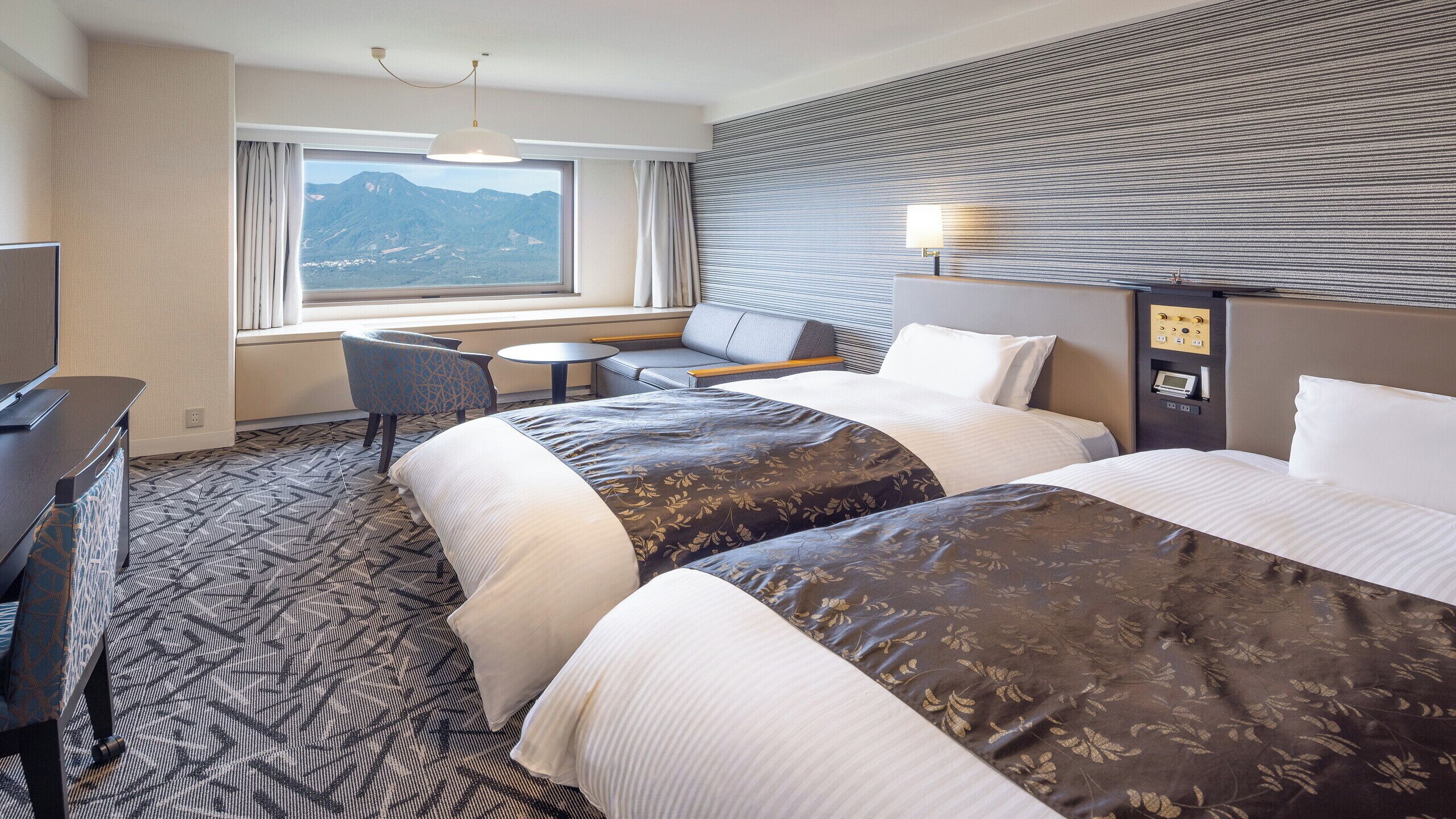 A hotel room with two beds, a desk, a chair, a small table, a TV, and a window with a mountain view.