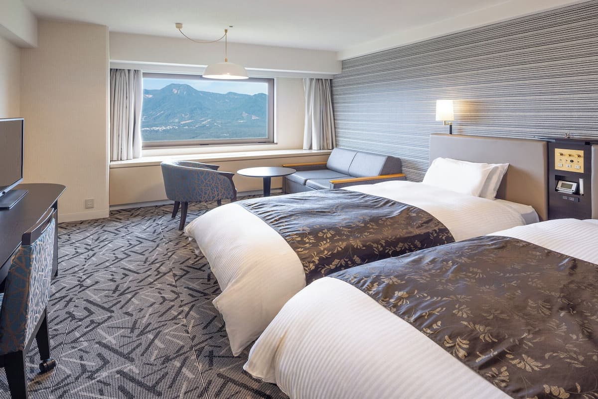 A hotel room with two beds, a desk, a chair, a small table, a TV, and a window with a mountain view.