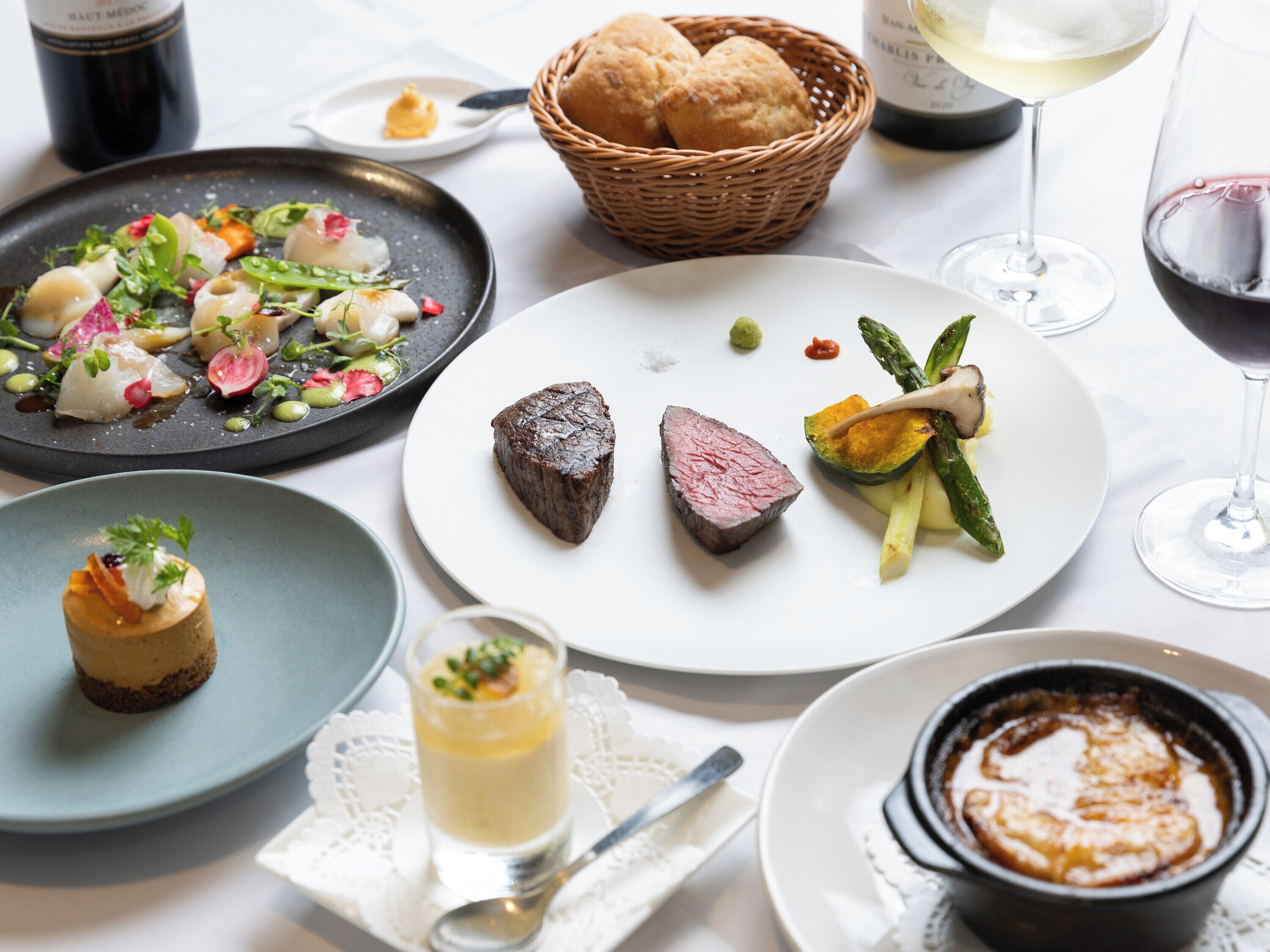 A variety of dishes on a table, including a steak, vegetables, and a dessert. There are also glasses of wine and a basket of bread.