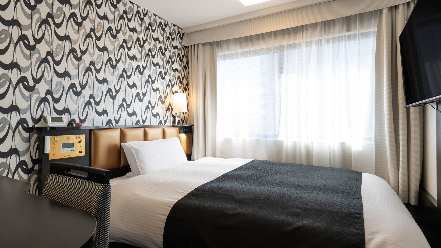A hotel room with a bed, a bedside table, a control panel, and a window with curtains.