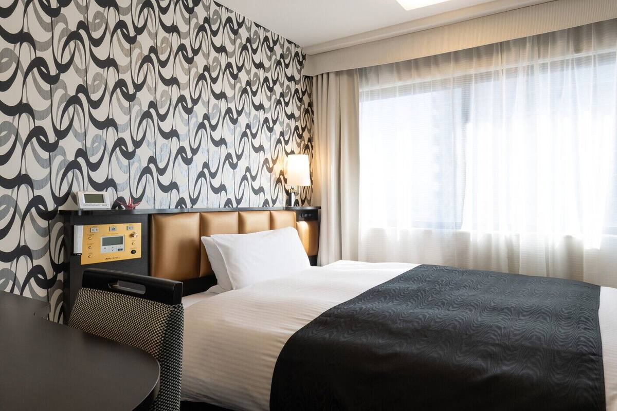 A hotel room with a bed, a bedside table, a control panel, and a window with curtains.