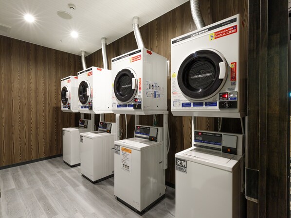 A row of front-loading washing machines in a commercial laundry room with wooden panel walls and a tiled floor. - APA Hotel Stay Toyama (Toyama)