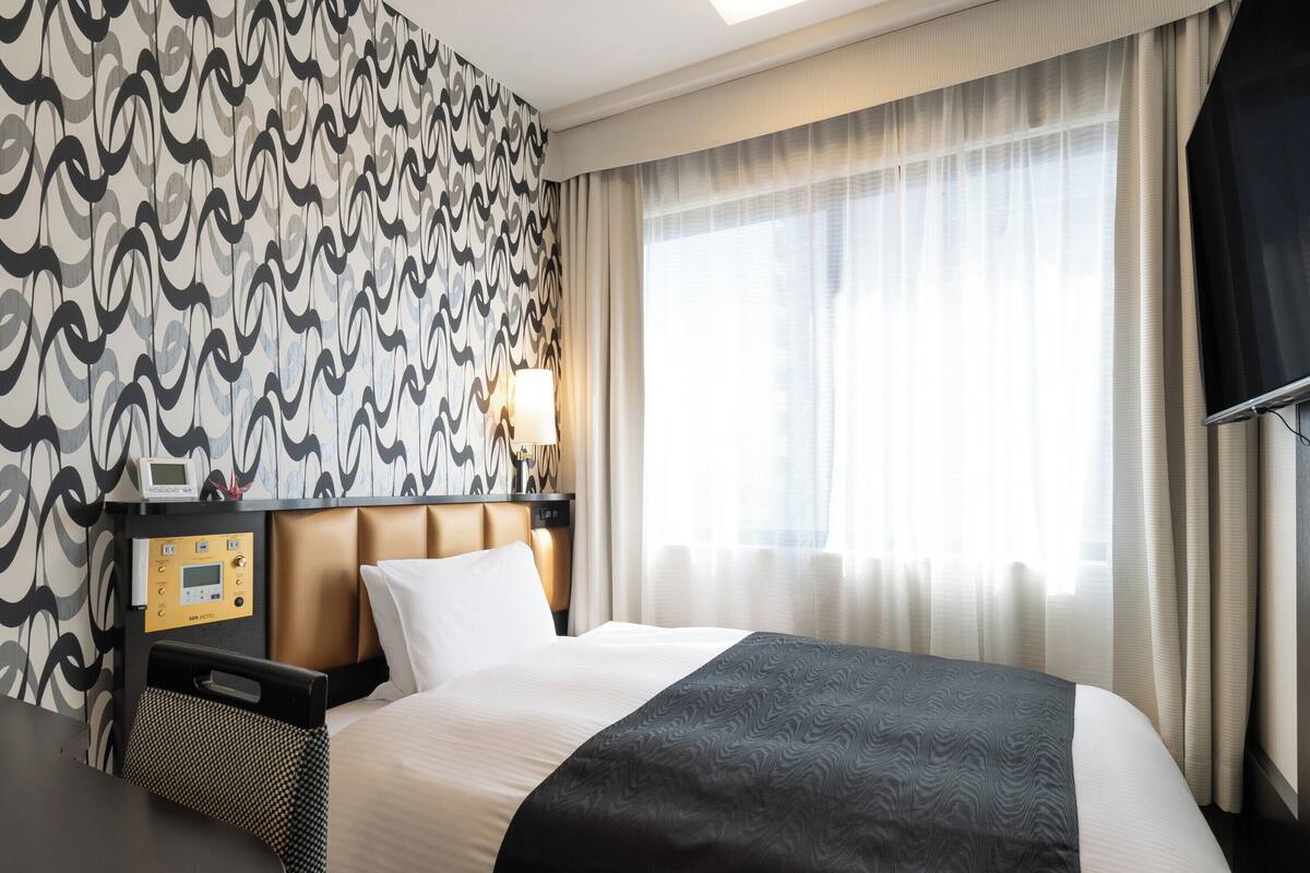 A hotel room with a large bed, a control panel, a desk, and a window with sheer curtains.