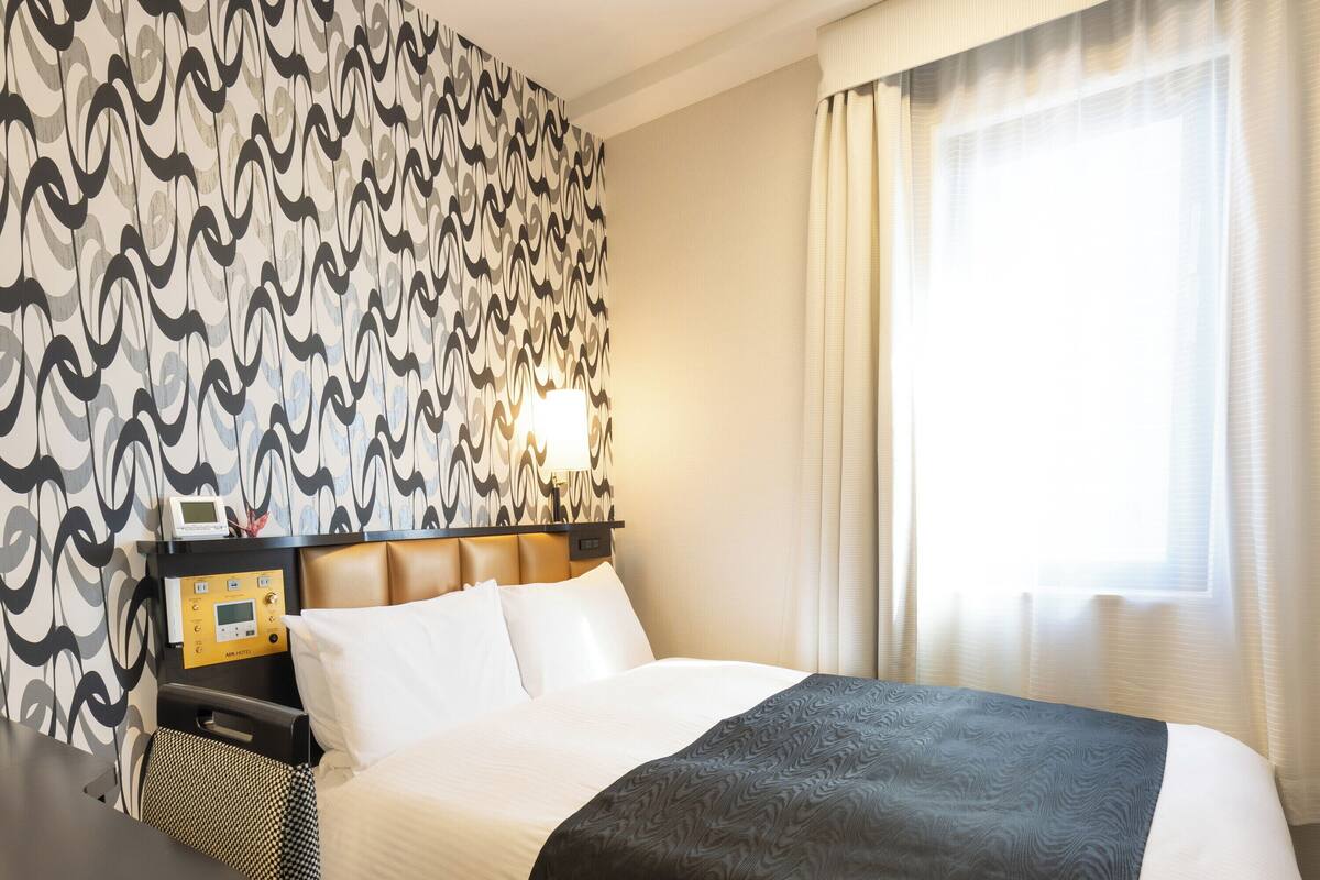 A hotel room with a patterned wallpaper, a bed with white and dark grey bedding, a bedside table with a lamp, and a window with sheer curtains.