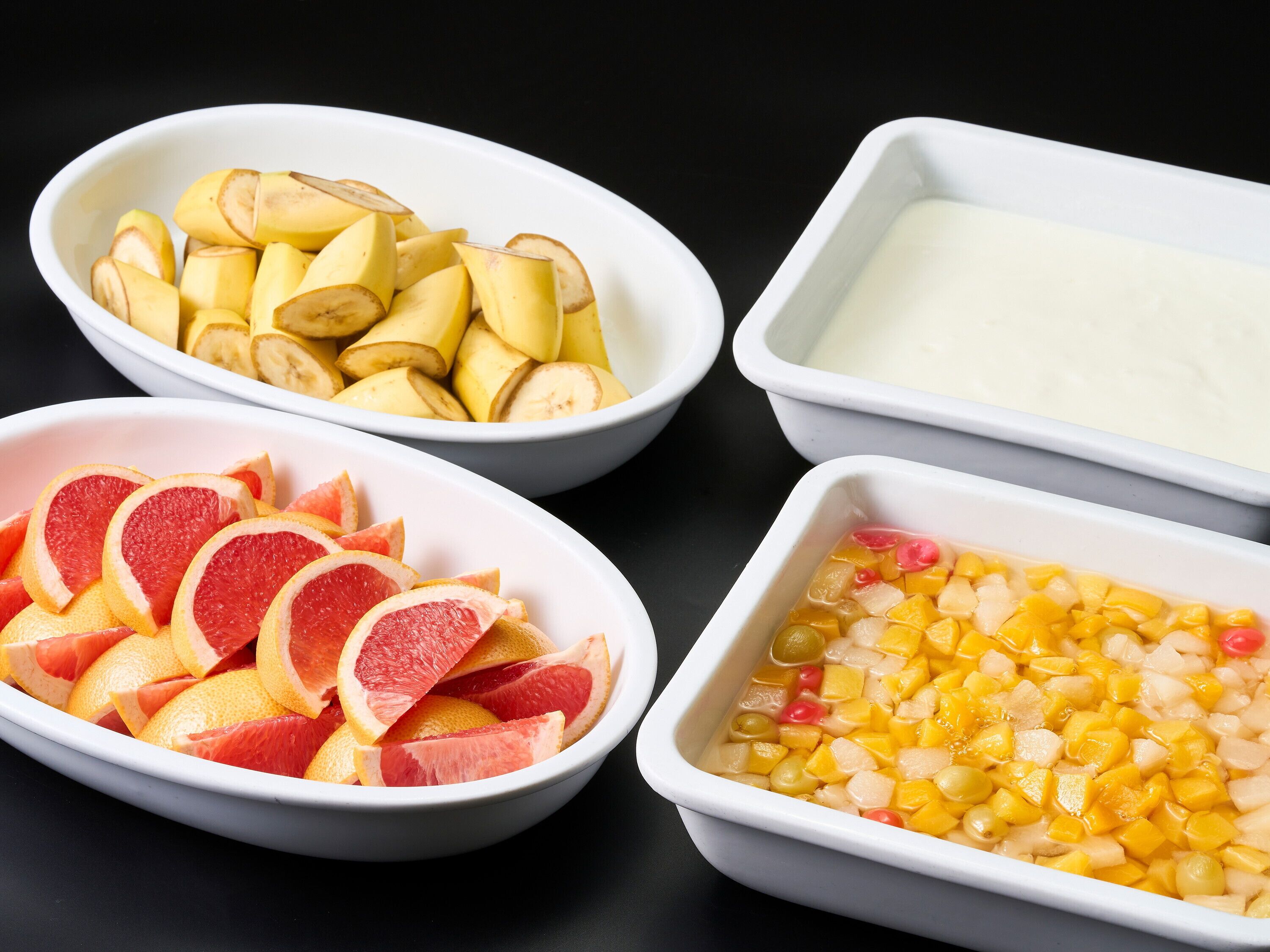 Three white bowls containing sliced fruits and a white rectangular dish with a yellowish liquid.