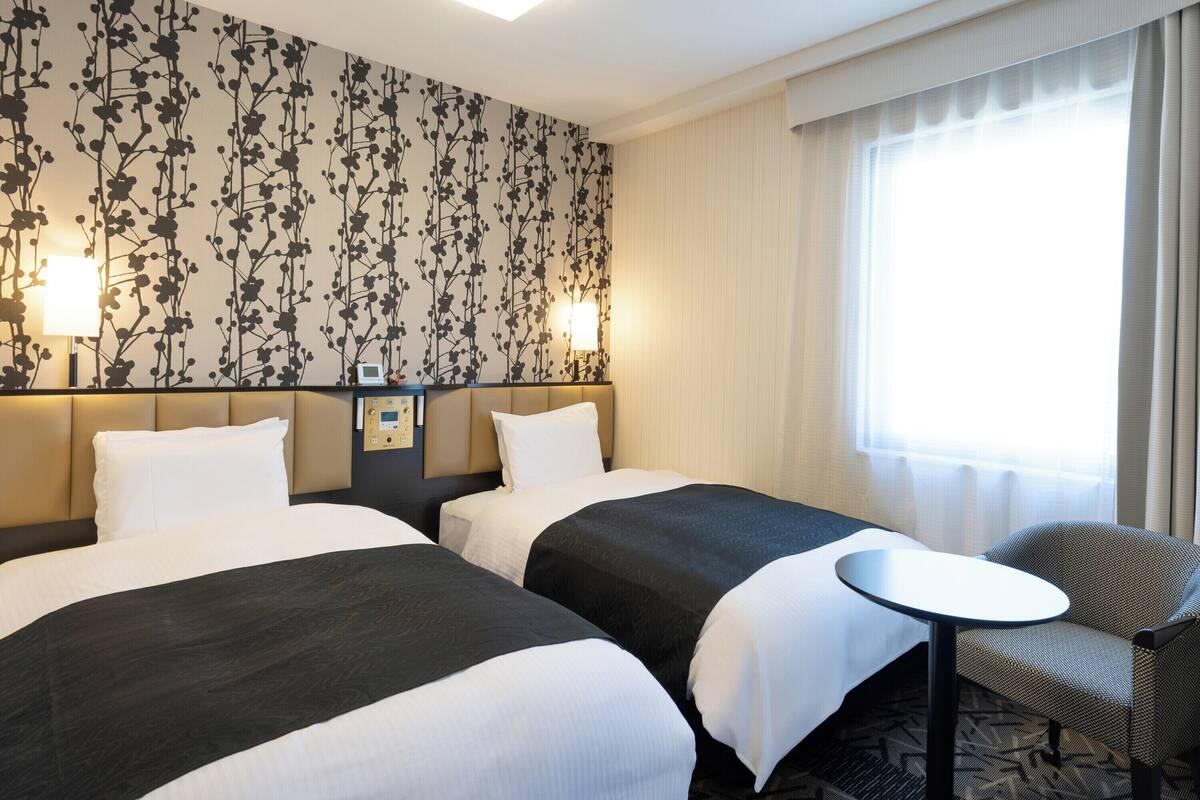 A hotel room with two beds, a small table, and a chair. There is a control panel on the wall between the beds.
