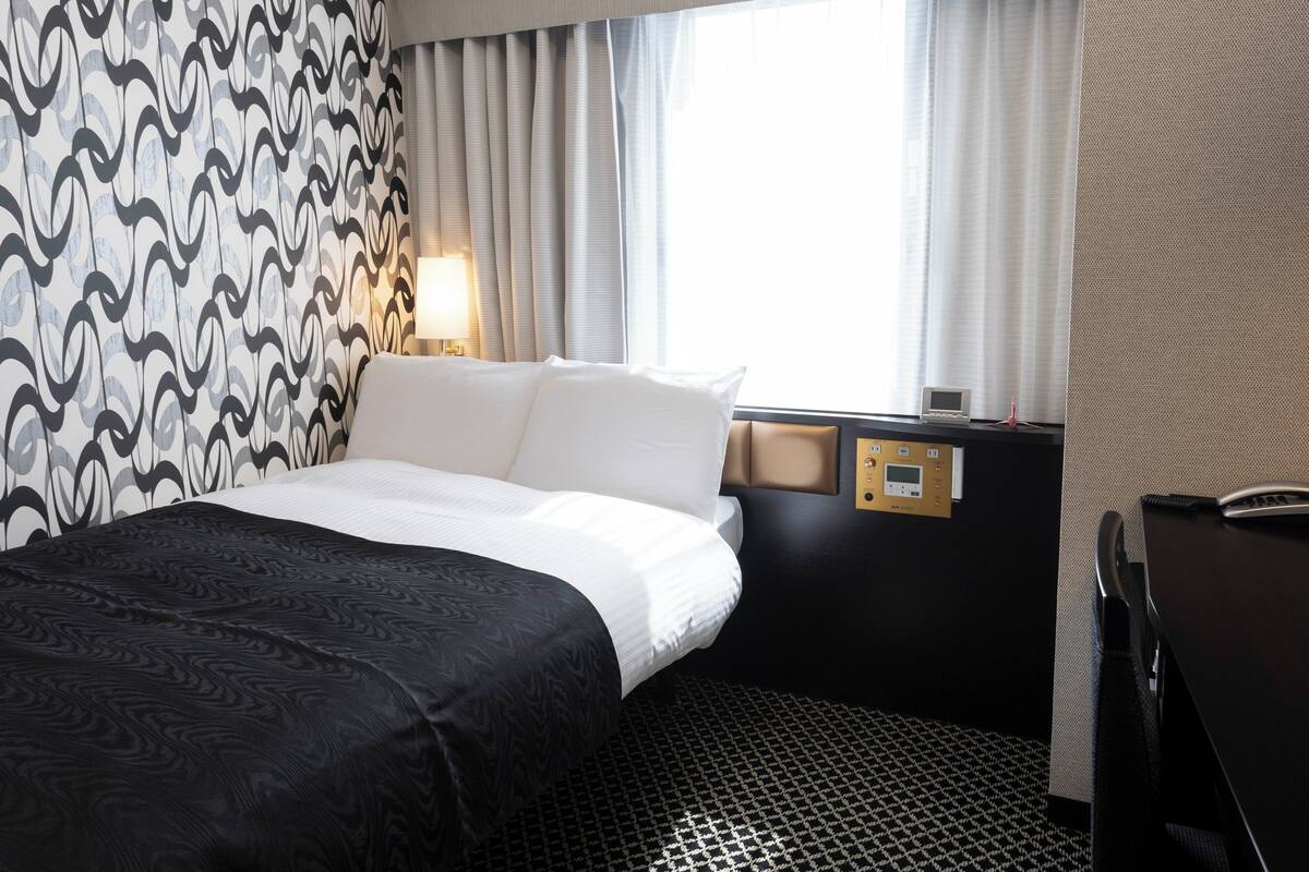 A hotel room with a bed, a desk, a control panel, and a window with curtains.