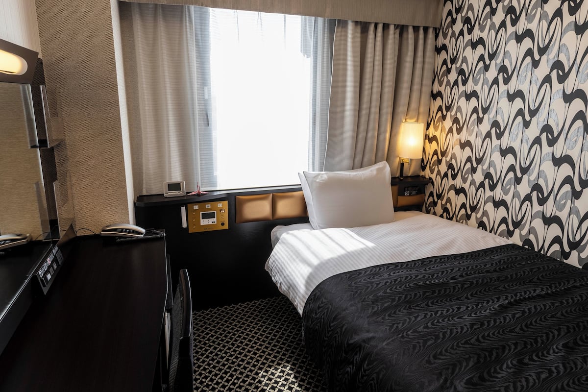 A hotel room with a bed, a desk with a telephone, a control panel, a lamp, and a window with curtains.