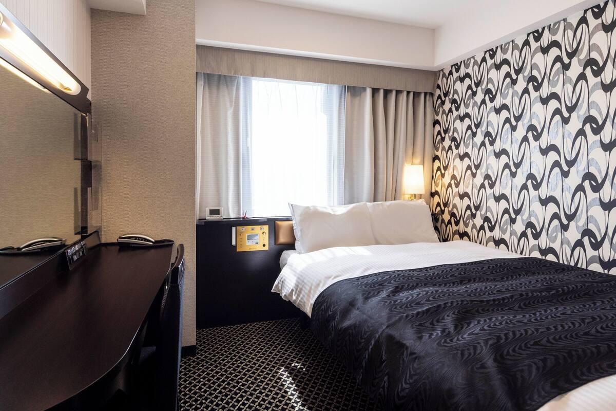 A hotel room with a bed, bedside table, lamp, and a window with curtains.