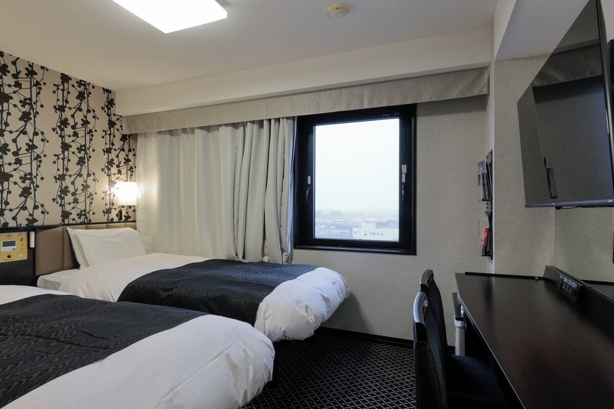 A hotel room with two beds, a large window with curtains, a flat-screen TV, and a wall-mounted control panel.