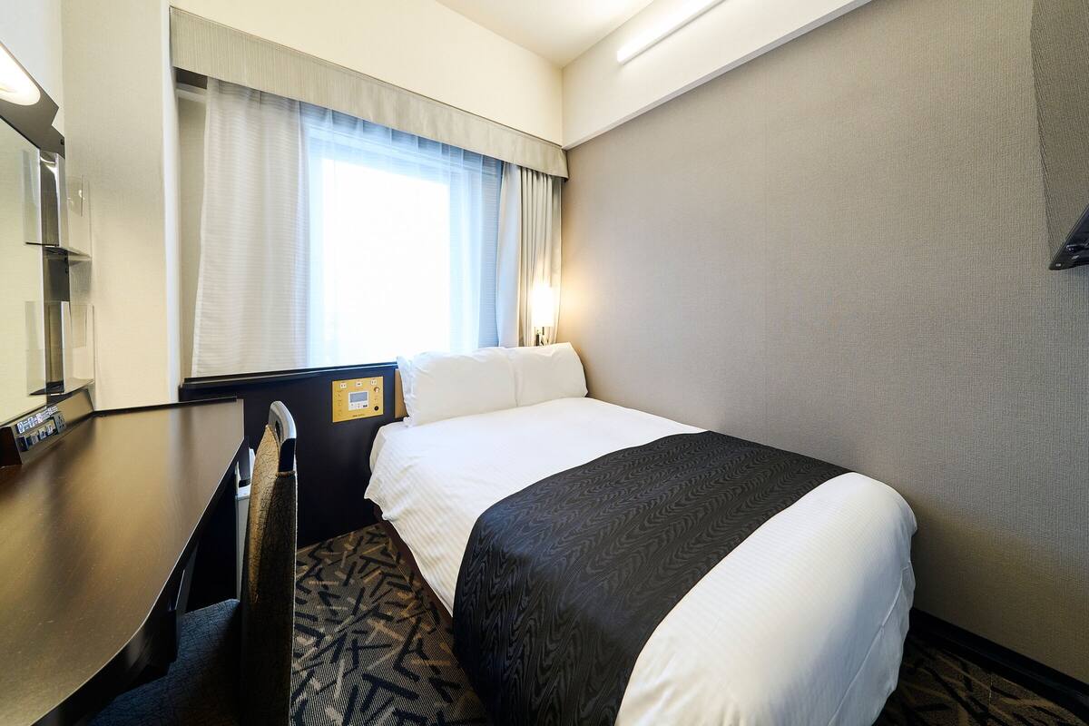 Hotelkamer met een bed, een bureau, een stoel, een raam met gordijnen en een bedieningspaneel aan de muur.