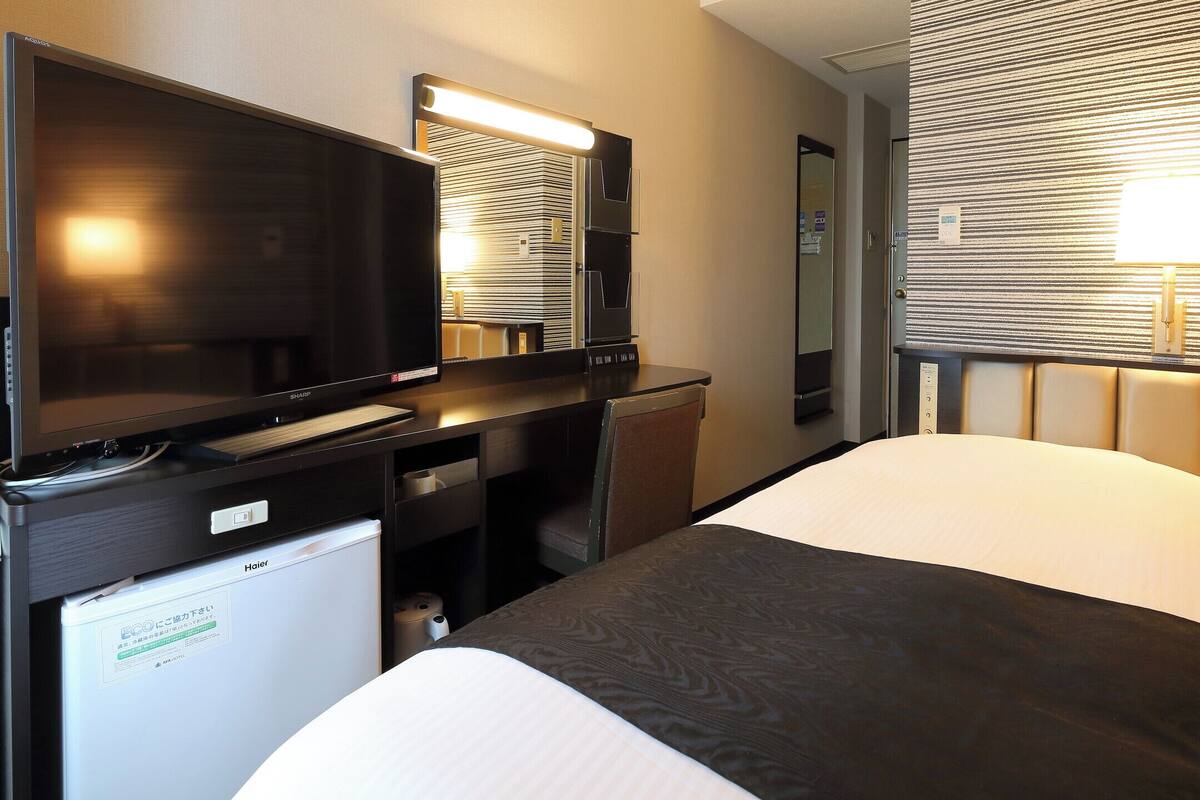 Een hotelkamer met een bed, een bureau met een televisie, een spiegel en een raam met jaloezieën.