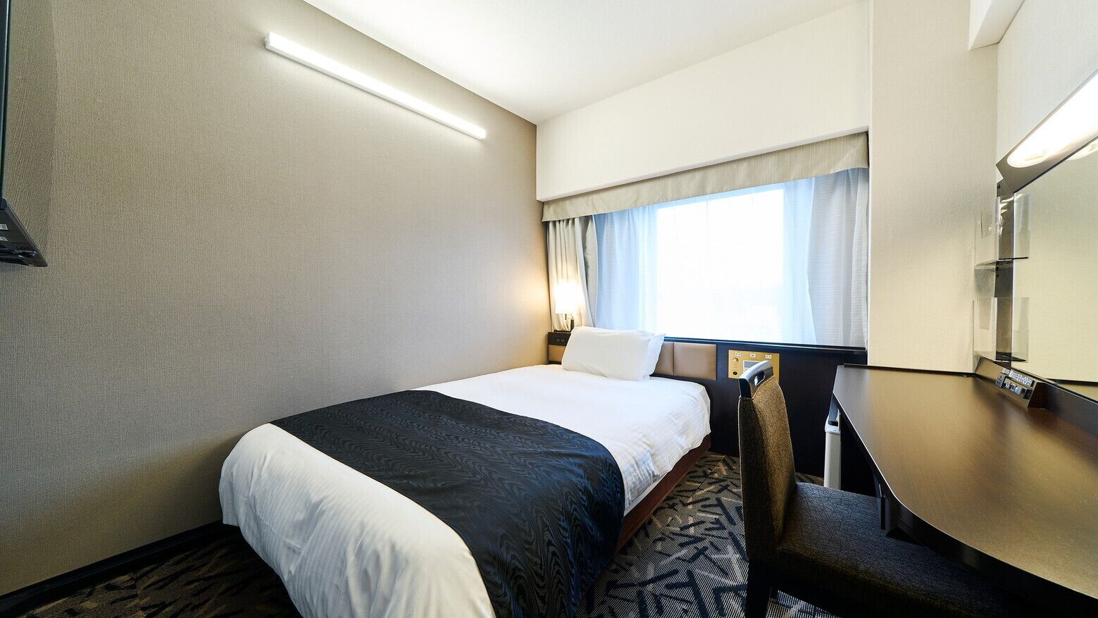 Une chambre d’hôtel équipée d’un lit, d’un bureau, d’une chaise, d’une fenêtre avec des rideaux et d’un téléviseur fixé au mur.