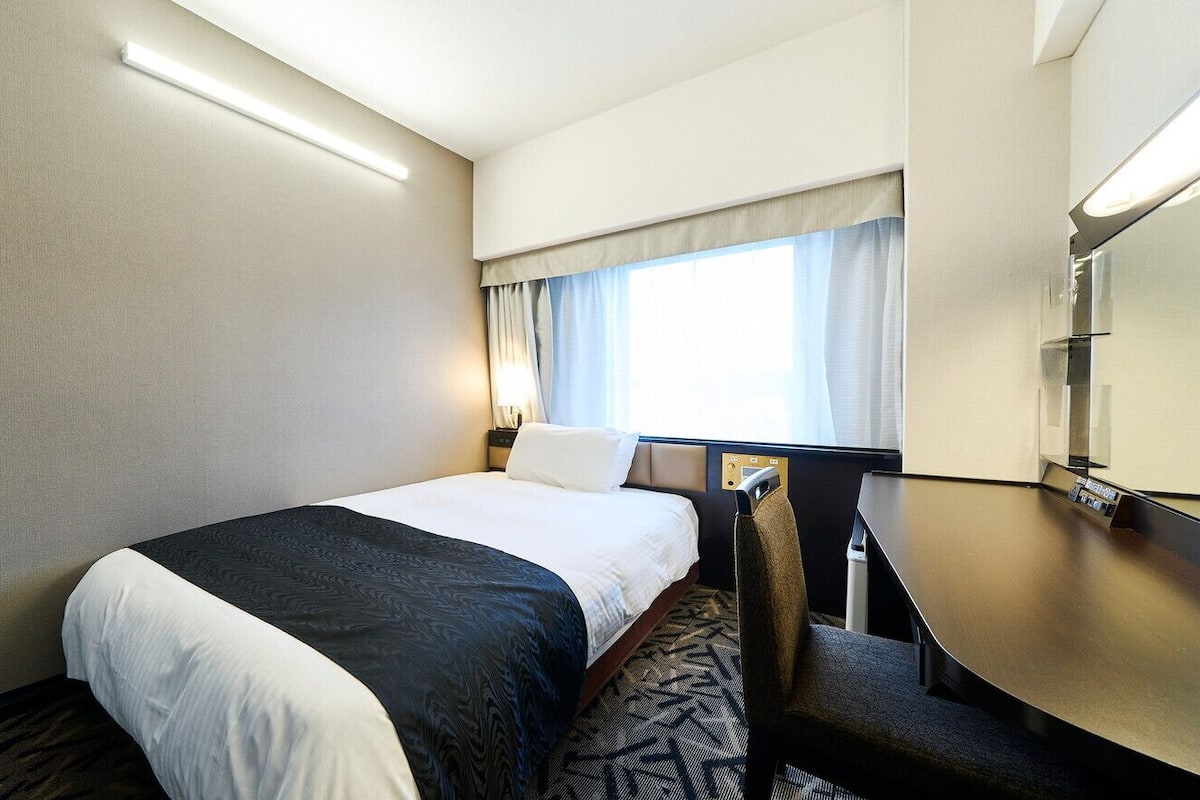 Une chambre d’hôtel équipée d’un lit, d’un bureau, d’une chaise, d’une fenêtre avec des rideaux et d’un téléviseur fixé au mur.