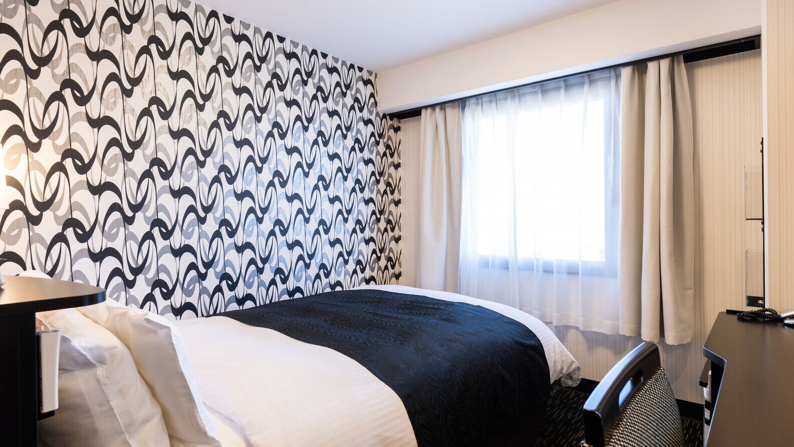 A hotel room with a patterned wall, a bed with a black and white bedspread, a desk with a lamp, and a window with sheer curtains.
