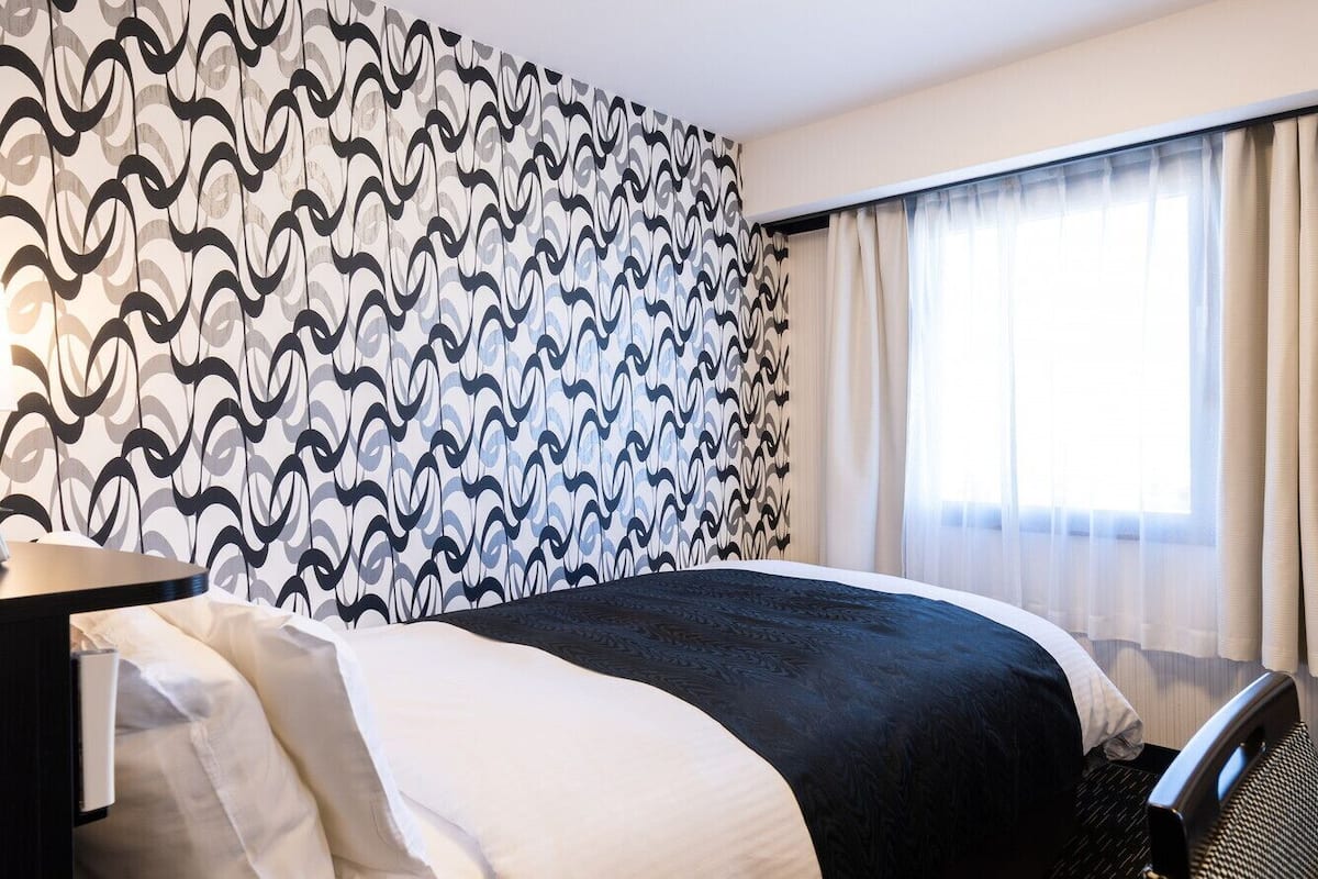 Ein Hotelzimmer mit Wandmuster, einem Bett mit schwarz-weiß gestreifter Bettdecke, einem Schreibtisch mit Lampe und einem Fenster mit durchsichtigen Vorhängen.
