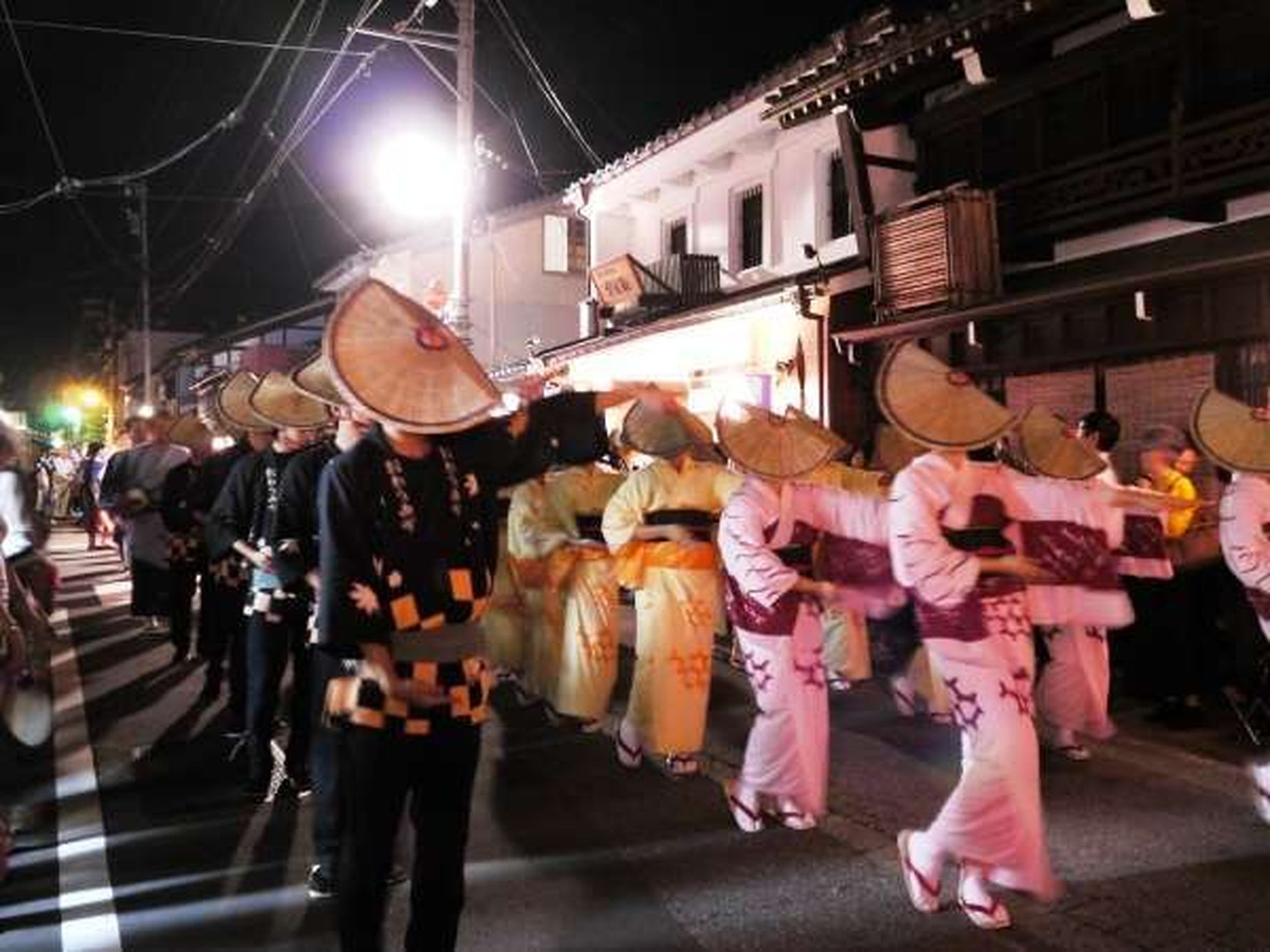 Escena callejera con gente vestida con trajes tradicionales, algunos usando sombreros cónicos, caminando por una calle de noche.