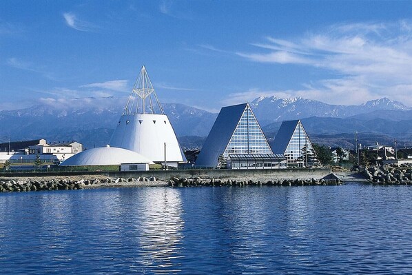 A modern architectural complex with a large white dome and two triangular buildings, situated by a body of water with mountains in the background. - APA Hotel Uozu Ekimae (Uozu)