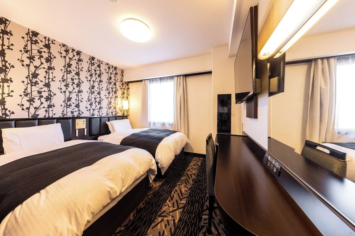 A hotel room with two beds, a desk, a flat-screen TV, and a large window with curtains.