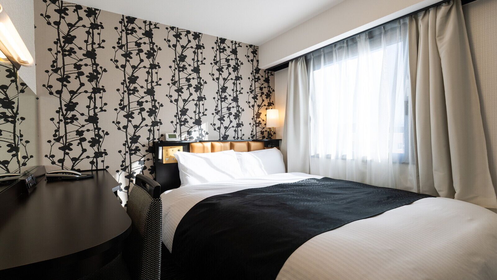 A hotel room with a bed, bedside table, lamp, and a wall with a floral pattern.