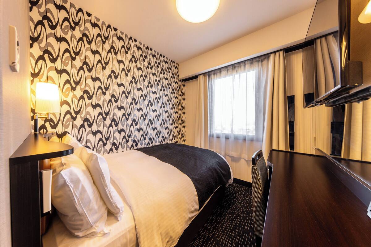 A hotel room with a bed, a desk, a lamp, a TV, and a window with curtains.