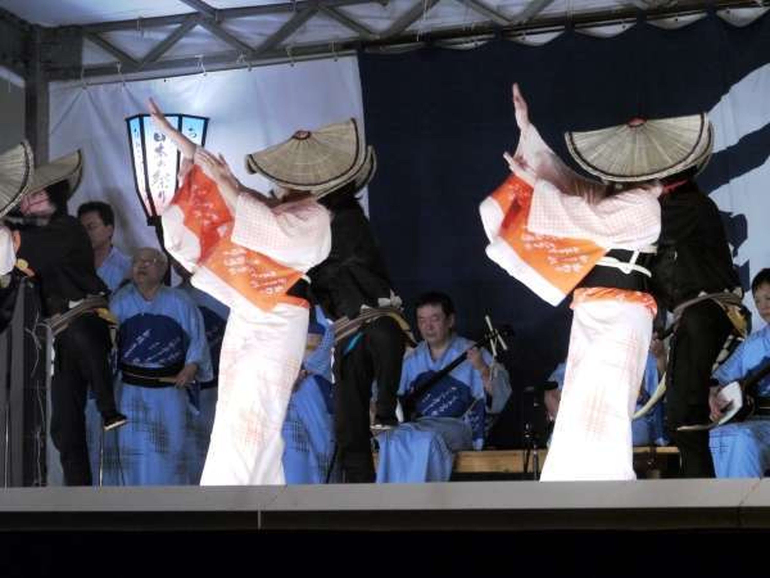 A stage with performers in traditional attire, a crowd of spectators, and a backdrop with Japanese characters.