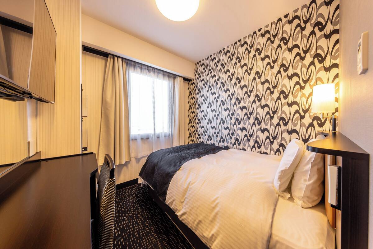A hotel room with a bed, a desk, a chair, a wardrobe, and a patterned wall.