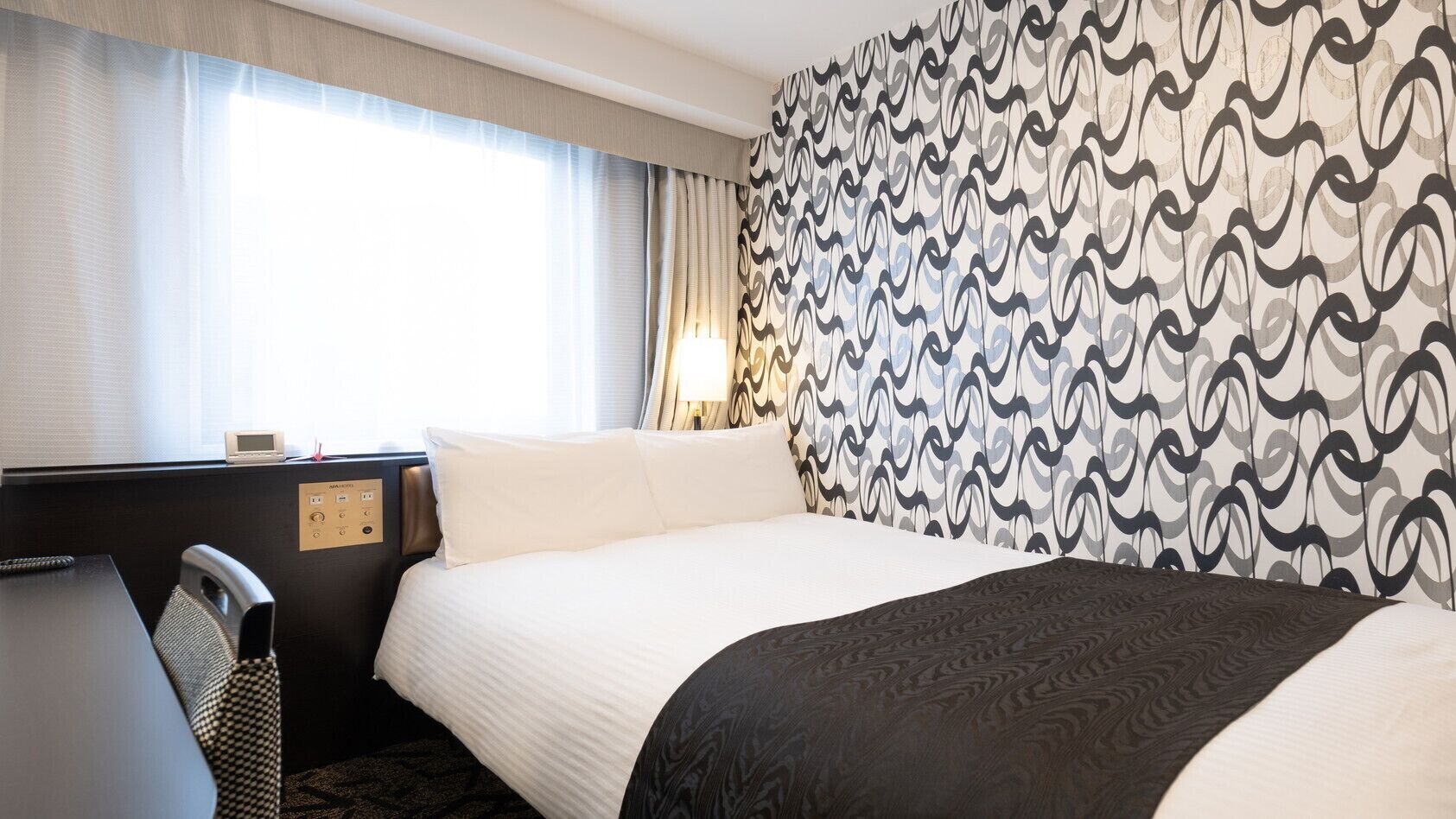 A hotel room with a bed, a desk, and a chair. There is a window with curtains, a wall with a patterned wallpaper, and a lamp on the desk.