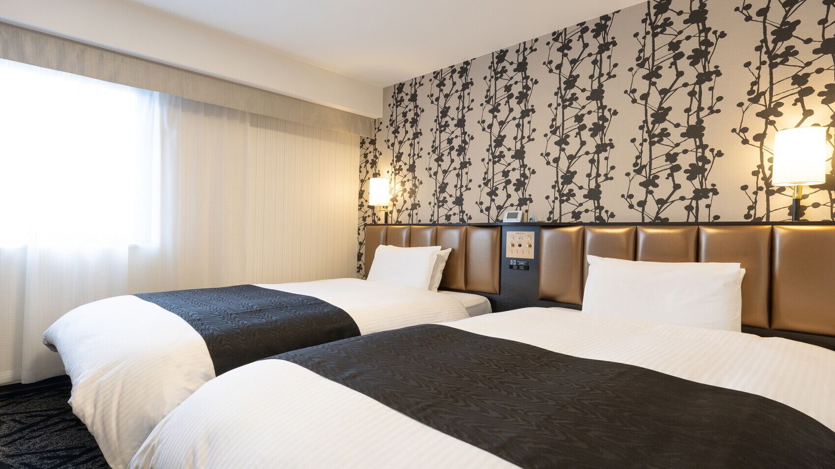 A hotel room with two beds, a headboard with controls, a window with curtains, and a wall with a floral pattern.