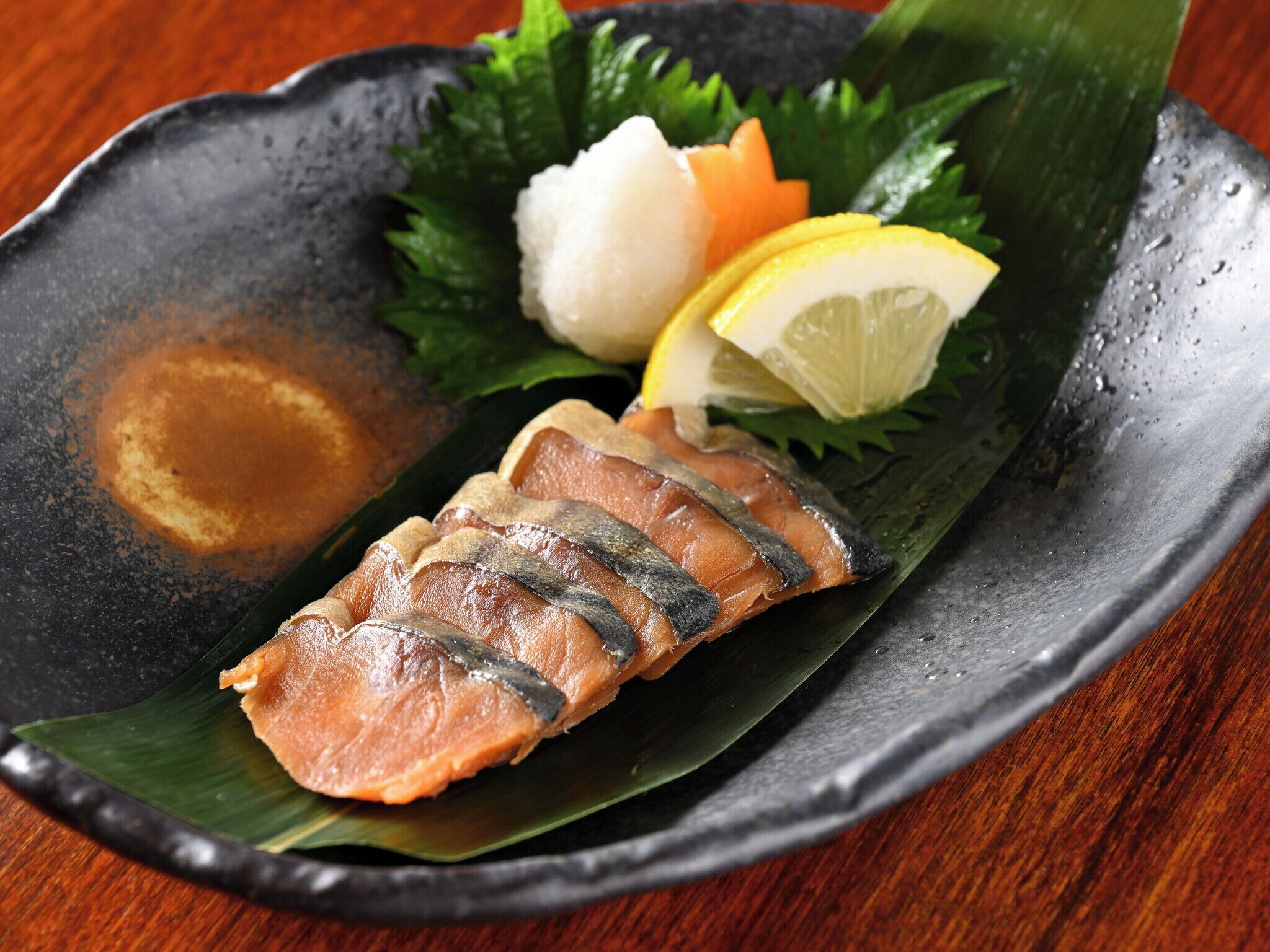 Un plato de cerámica negra con un trozo de salmón, un bollo blanco y una cuña de limón sobre una mesa de madera.