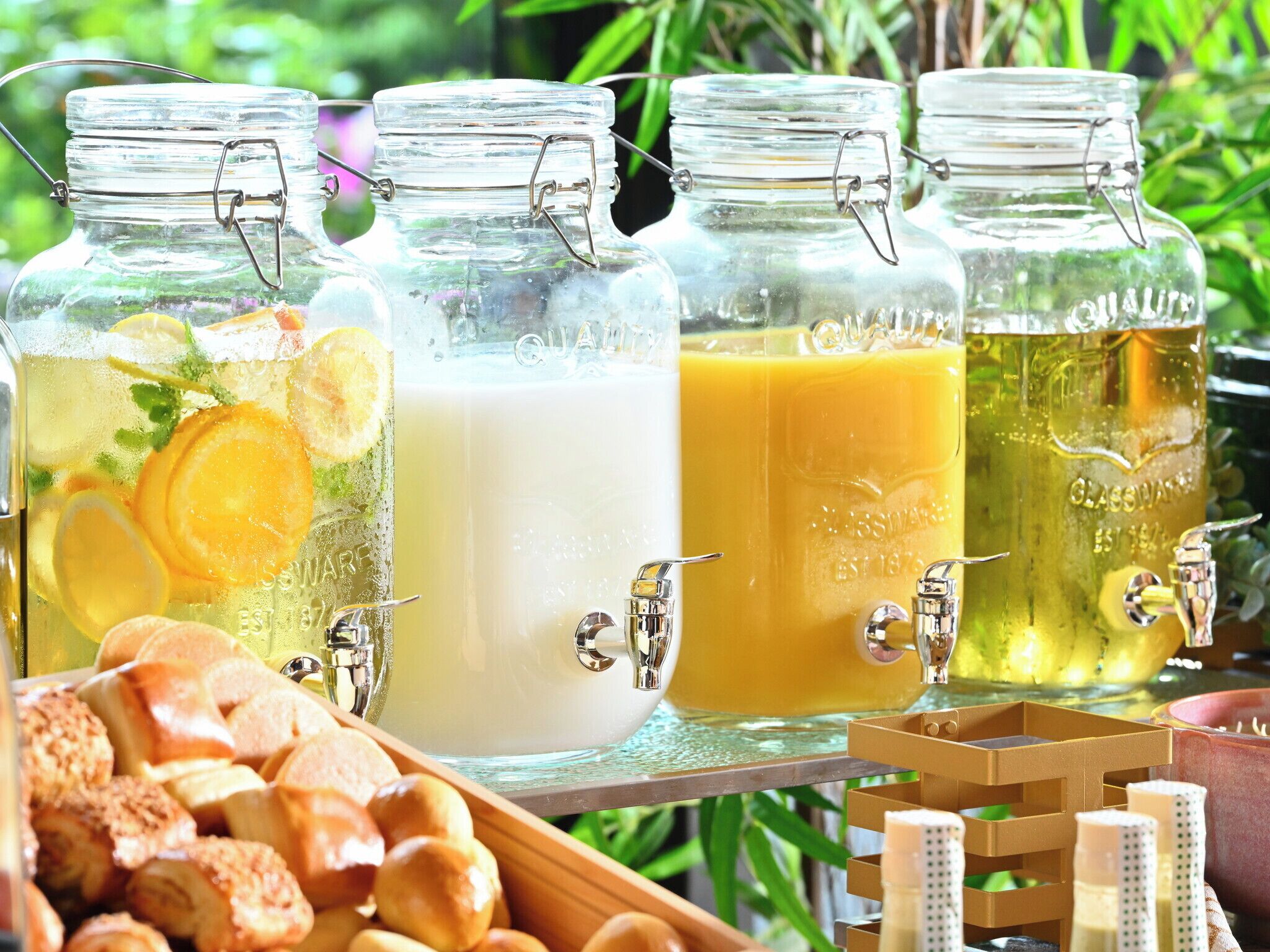 Four glass jars with metal clasps, each containing a different beverage and garnished with citrus slices. A tray with bread rolls is in the foreground.