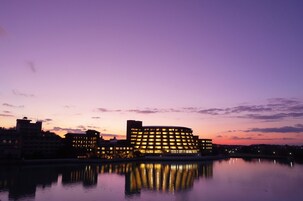 A waterfront hotel with multiple buildings, a calm body of water reflecting the structures, and a clear sky.