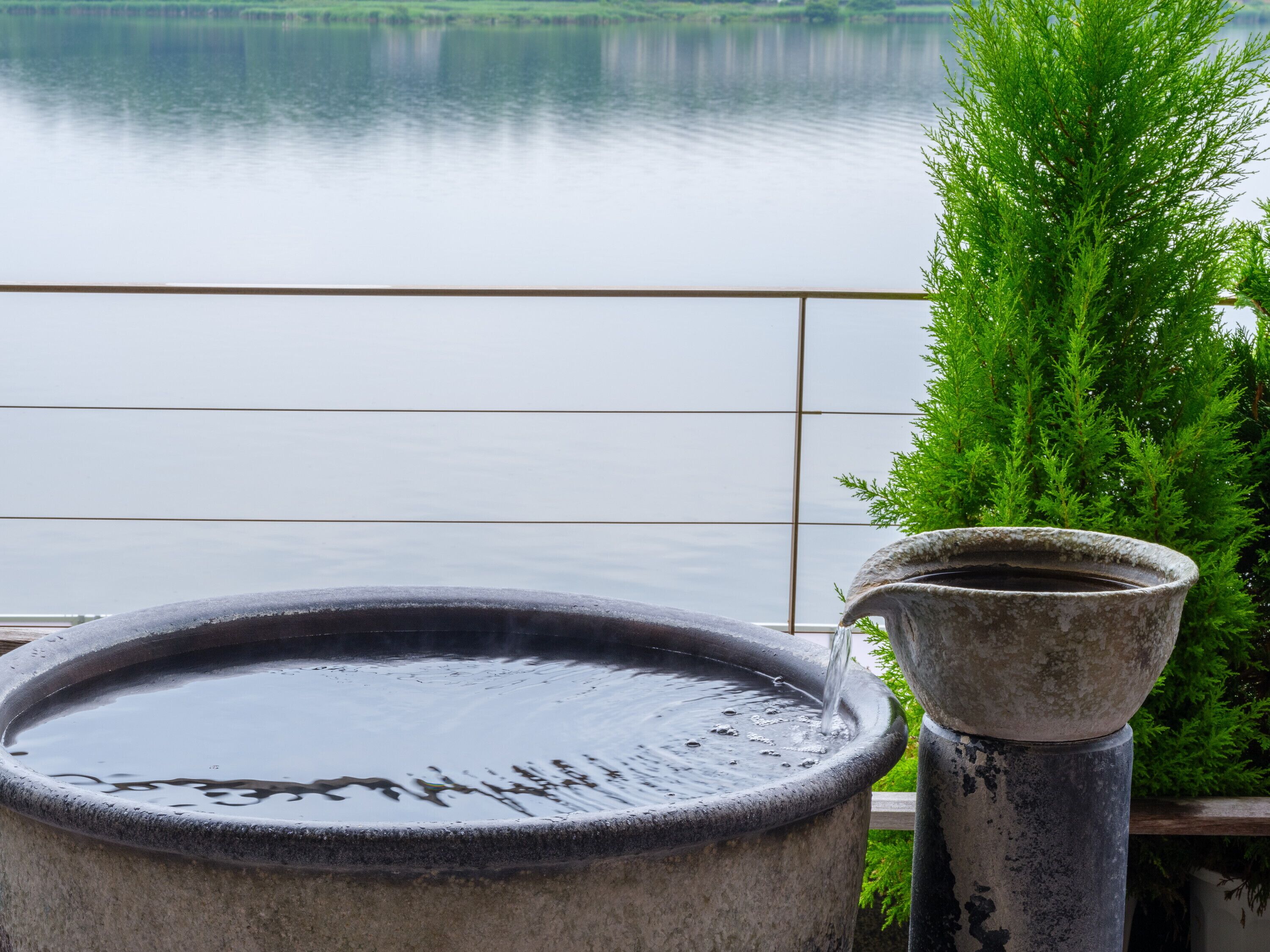A stone water feature with a spout, set against a backdrop of a calm lake and a green hedge.